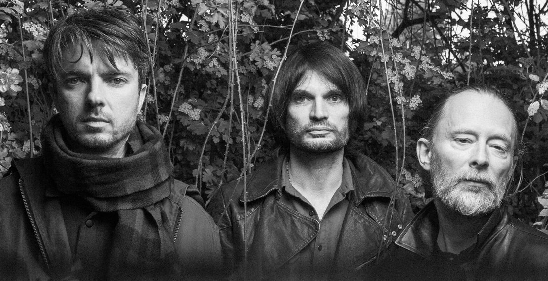 a black and white photo of three men with long hair and beards, standing in front of a stand of trees